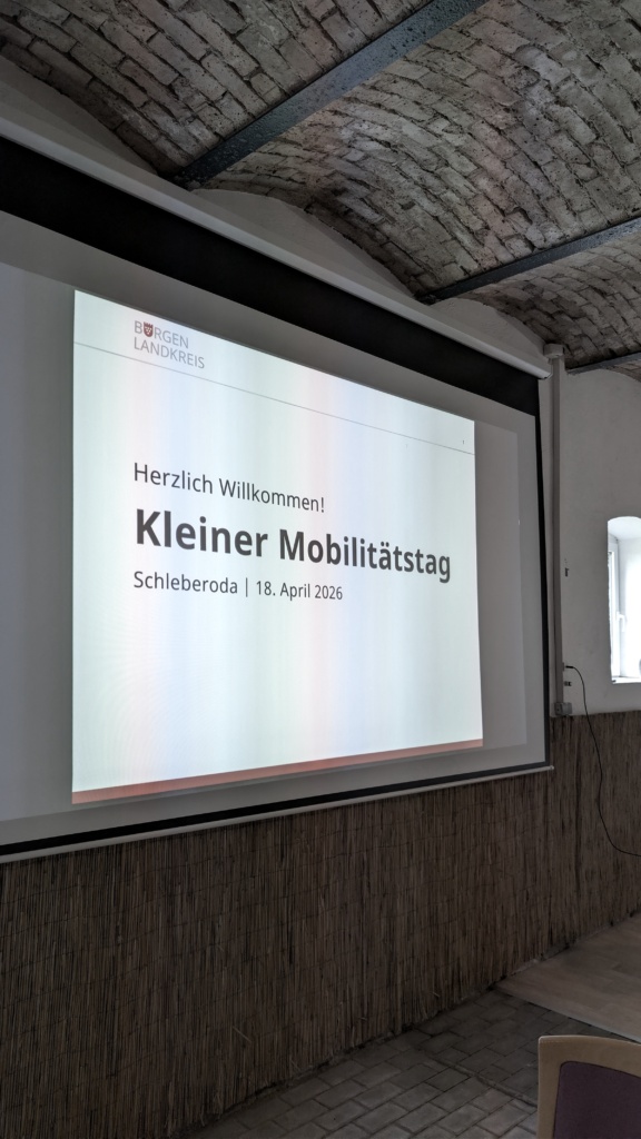 Eine Leinwand in einem Raum mit Ziegelgewölbedecke. Auf der Leinwand steht „Herzlich Willkommen! Kleiner Mobilitätstag – Schleberoda, 18. April 2026“ mit dem Wappen des Burgenlandkreises.