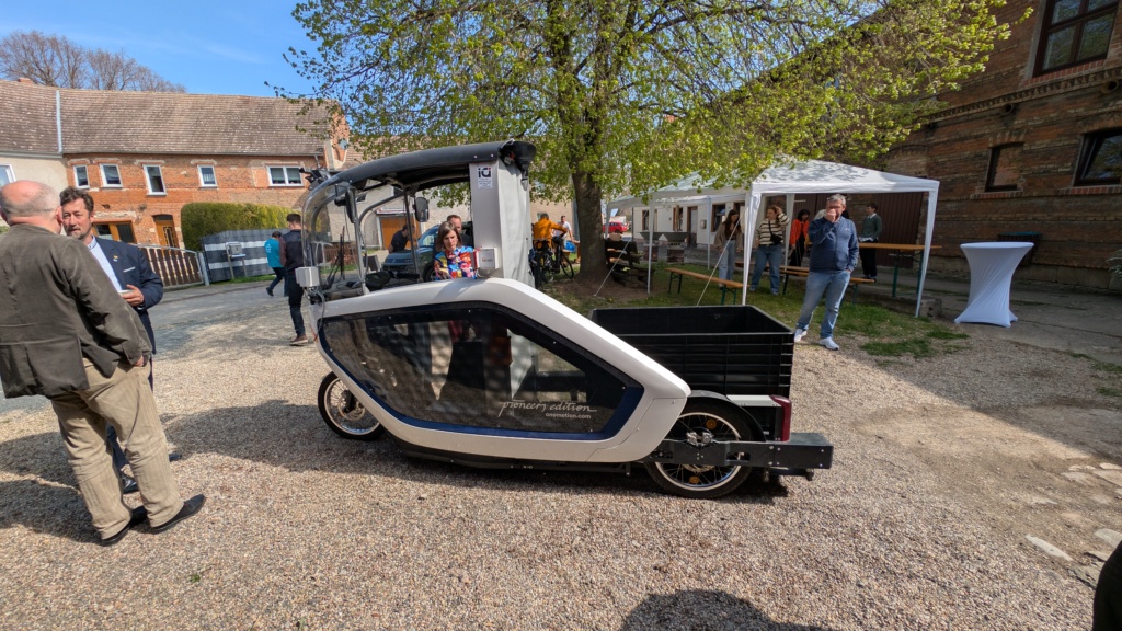 Ein weiß-schwarzes, geschlossenes, vierrädriges E-Lastenrad der Marke „Pioneer Edition“ steht im Hof des Veranstaltungsorts. Im Hintergrund ein Pavillon mit Stehtischen, Ziegelfassaden und Besucherinnen und Besucher im Gespräch.