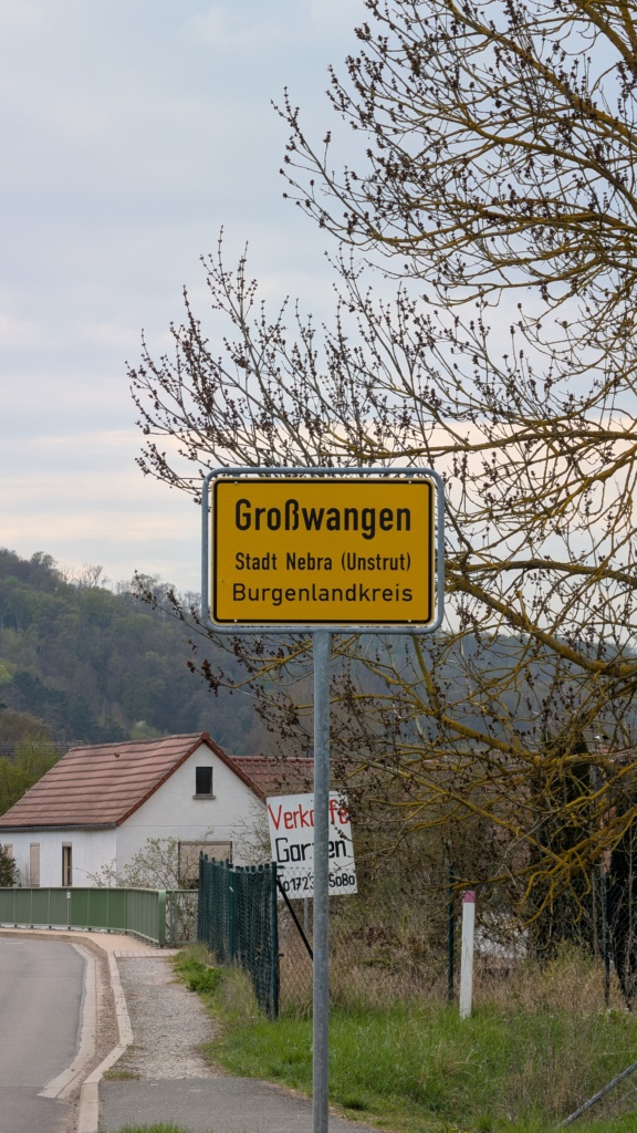 Ein gelbes Ortseingangsschild mit der Aufschrift „Großwangen, Stadt Nebra (Unstrut), Burgenlandkreis“ vor blassblauem Himmel. Im Hintergrund ein weißes Haus mit rotem Ziegeldach, Sträucher und kahle Bäume im zeitigen Frühjahr.