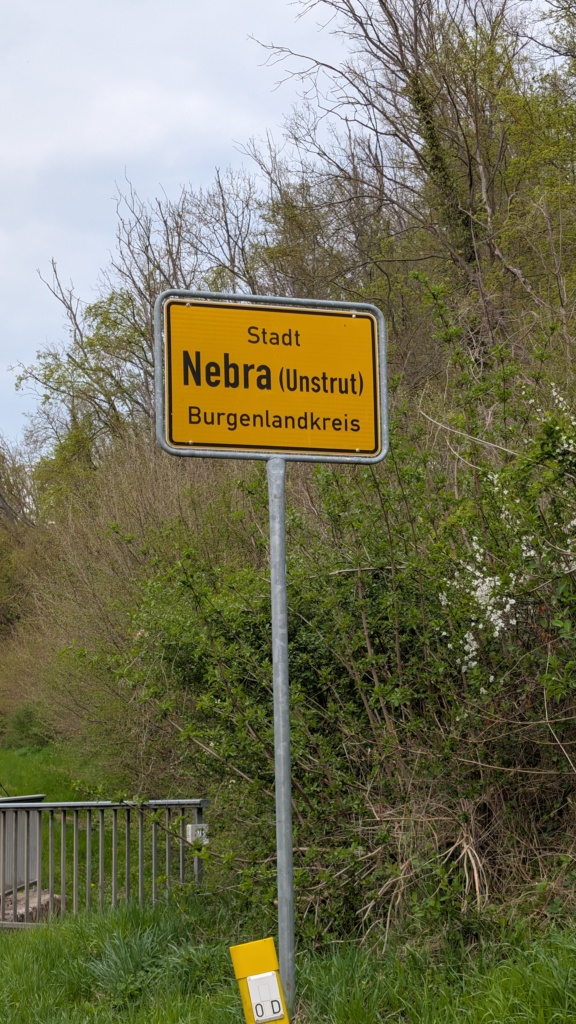Gelbes Ortseingangsschild mit der Aufschrift „Stadt Nebra (Unstrut), Burgenlandkreis“. Im Hintergrund Sträucher und Gehölz, am rechten Bildrand weiße Blüten eines Schlehdorns.