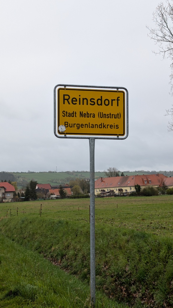 Gelbes Ortseingangsschild mit der Aufschrift „Reinsdorf, Stadt Nebra (Unstrut), Burgenlandkreis“ vor grauem, bedecktem Himmel. Im Hintergrund eine Reihe von Häusern mit roten Ziegeldächern und bewirtschaftete Felder.