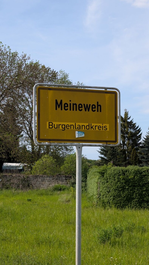 Gelbes Ortseingangsschild „Meineweh, Burgenlandkreis“ vor bewölktem, hellem Himmel. Auf dem Schild klebt ein kleiner Aufkleber. Im Hintergrund eine Bruchsteinmauer, Sträucher und Nadelbäume.