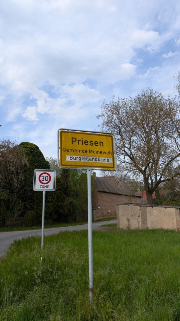 Gelbes Ortseingangsschild „Priesen, Gemeinde Meineweh, Burgenlandkreis“ vor leicht bewölktem Himmel. Daneben ein „Tempo-30-Zone“-Schild. Im Hintergrund ein Backsteingebäude mit niedriger Mauer und ein knorriger, noch unbelaubter Baum.