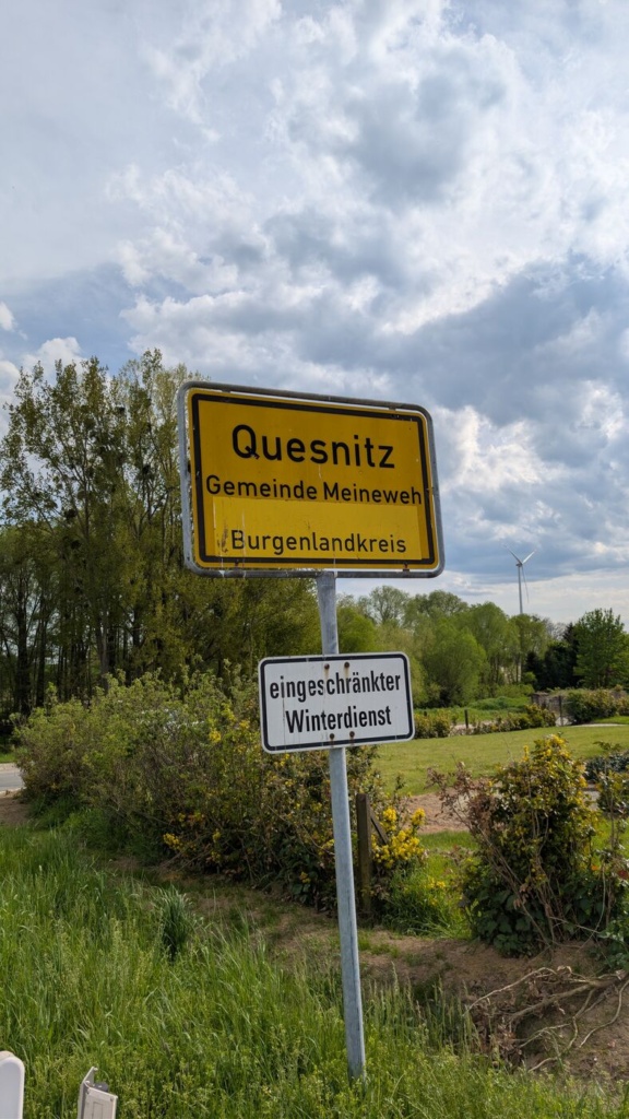 Ein gelbes Ortseingangsschild mit der Aufschrift „Quesnitz, Gemeinde Meineweh, Burgenlandkreis“. Im Hintergrund ein bewölkter Himmel, Bäume und ein einzelnes Windrad. Unter dem Ortsschild ein kleineres weißes Schild mit der Aufschrift „eingeschränkter Winterdienst“.