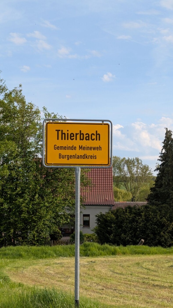 Gelbes Ortseingangsschild „Thierbach, Gemeinde Meineweh, Burgenlandkreis“ vor blauem Himmel mit Schäfchenwolken. Im Hintergrund ein einzelnes Wohnhaus mit rotem Ziegeldach, links und rechts Bäume.
